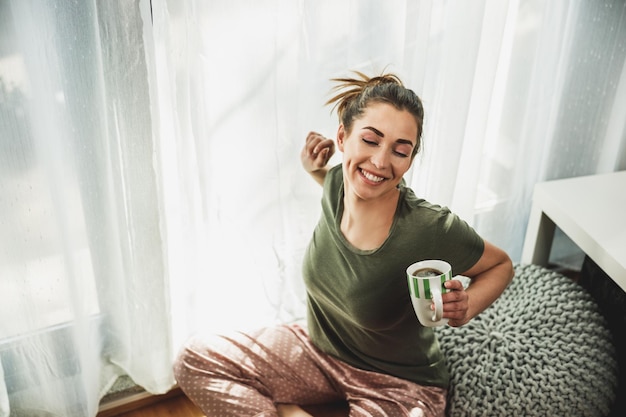 Young smiling woman enjoying coffee in the cozy morning\
sunshine near the window at home or hotel room.