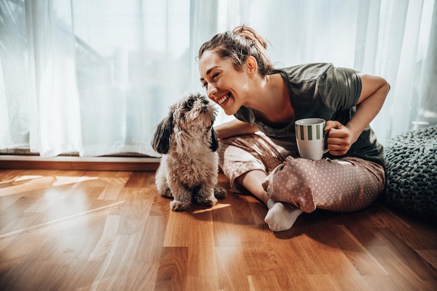 自宅で居心地の良い朝に彼女のシーズー犬とコーヒーを楽しんでいる若い笑顔の女性。