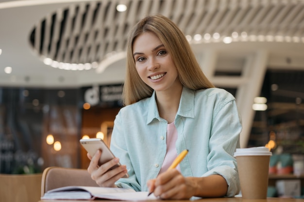 Young smiling woman freelancer  using mobile phone, taking tones, working in cafe