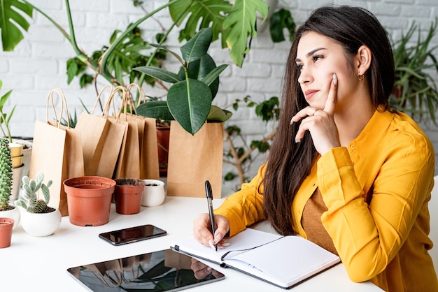 Young smiling woman gardener making notes in the notepad surrounded with plants