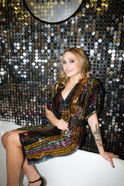 Young smiling woman in glamorous dress holding flute of champagne while sitting on bathtub by glittering wall