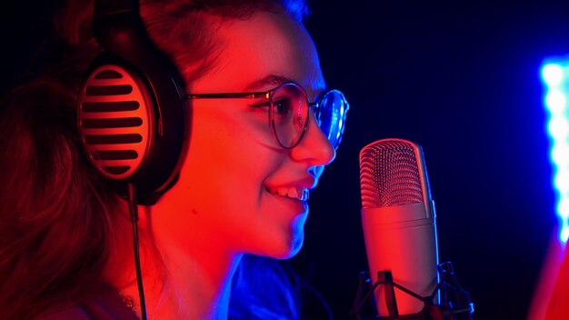 A young smiling woman in glasses singing by the microphone in neon lighting in the studio