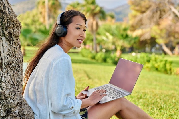 Young smiling woman in headphones using laptop sitting in park on grass