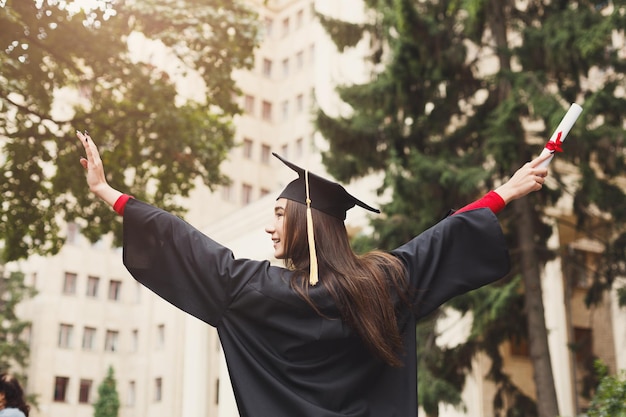 卒業証書を保持している大学の彼女の卒業式の日に若い笑顔の女性。教育、資格およびガウンの概念。