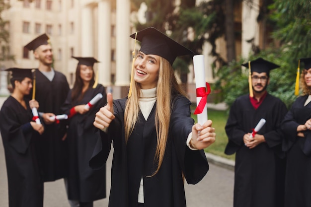 大学の卒業式の日に、学生の多民族のグループと一緒に立っている若い笑顔の女性。教育、資格およびガウンの概念。