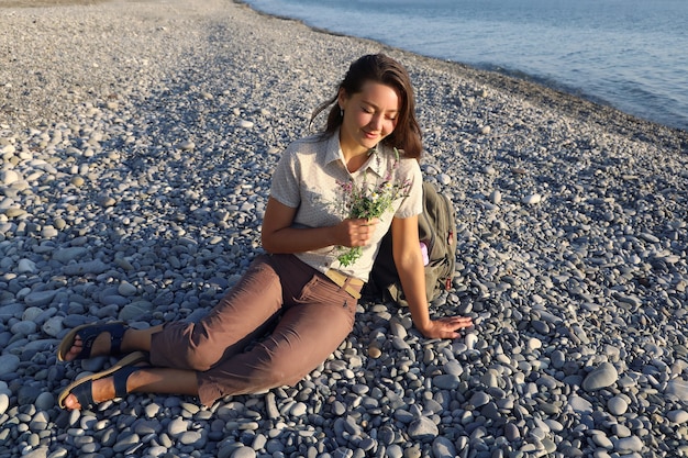 Giovane donna escursionista sorridente si siede sulla spiaggia di ciottoli deserta e detiene un piccolo mazzo di fiori di campo