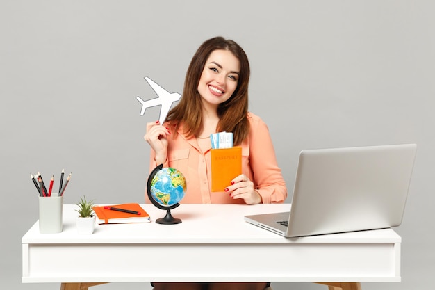 Young smiling woman holding paper air plane, Earth world globe, passport boarding pass ticket work at desk with pc laptop isolated on gray background. Achievement business career lifestyle concept.