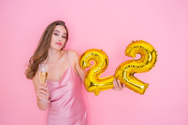 Young smiling woman holds gold foil balloon and glass of champagne on pink background birthday party