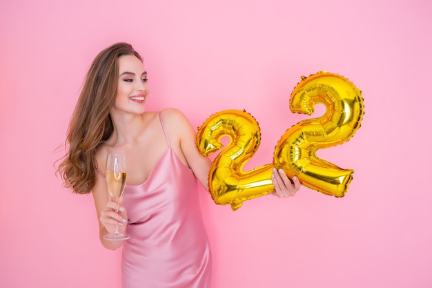 Young smiling woman holds gold foil balloon and glass of champagne on pink background birthday party