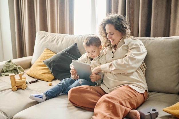 写真 息子が持っているタブレットの画面を指しているカジュアルウェアの若い笑顔の女性