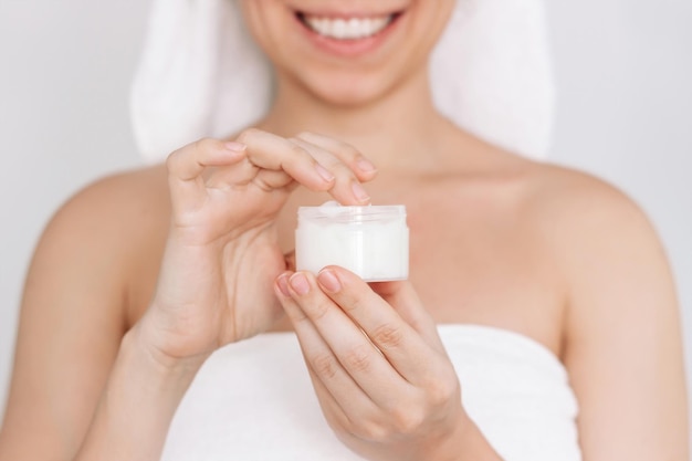 写真 シャワーの後、瓶を持った手で保湿クリームを取る白いタオルを着た若い笑顔の女性