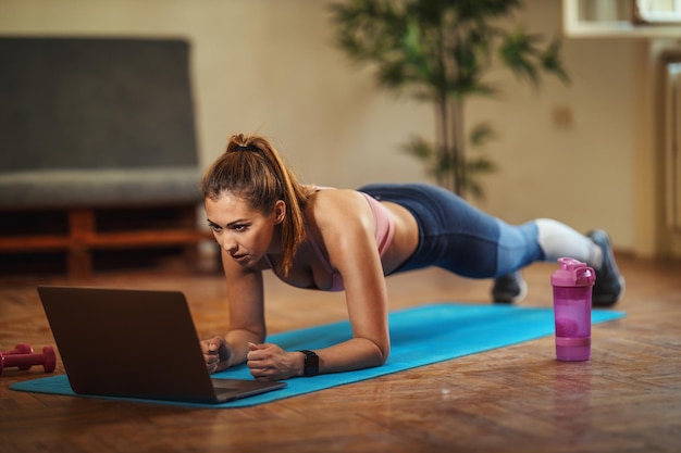 Young smiling woman is doing plank exercises in the living room\
on floor mat at home, looking at the laptop, in morning\
sunshine.