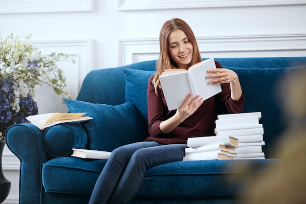 Young smiling woman is reading on a sofa with a pile of books