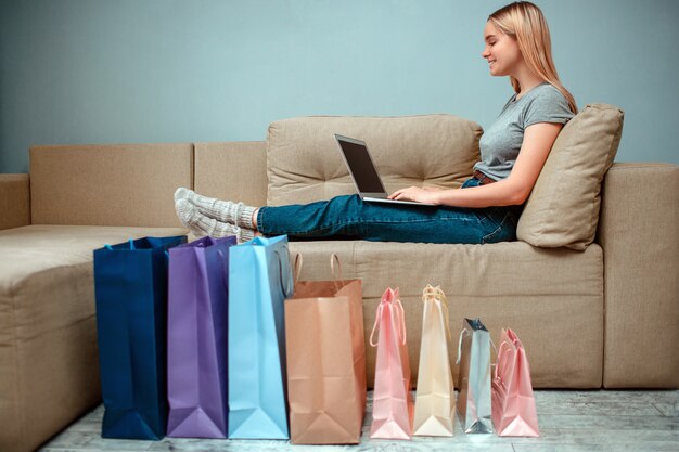 Young smiling woman is sitting on sofa and using laptop near colorful shopping bags