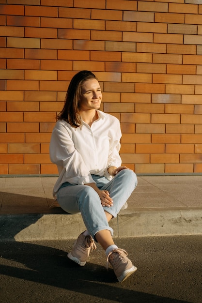 Young smiling woman is sitting in the sunlight outdoors near a brick wall