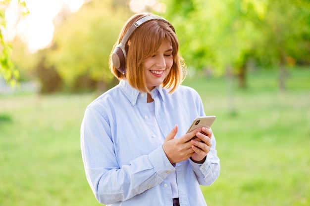 Young smiling woman listening music with wireless headphones at sunset and typing message on phone