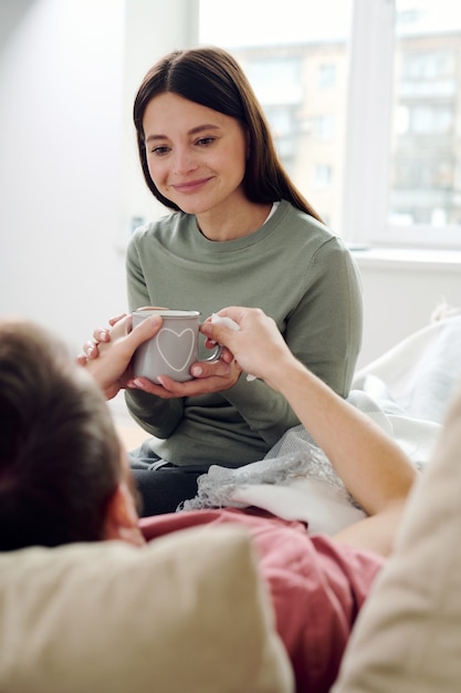 Giovane donna sorridente che guarda il marito malato sdraiato sul divano mentre gli dà una bevanda calda, gli tocca la mano e sorride contro la finestra