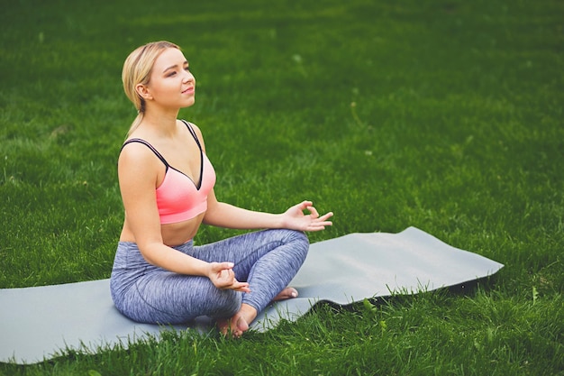 Young smiling woman outdoors, meditation exercises. Girl does lotus pose for relaxation. Wellness, calmness, relax, healthy, active lifestyle concept