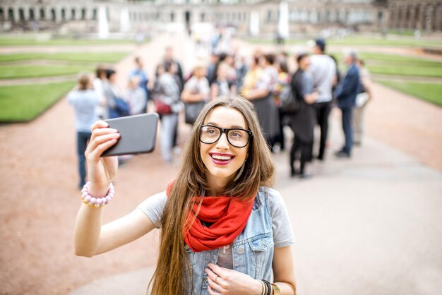 Молодая улыбающаяся женщина фотографирует со смартфоном во время посещения с туристической группой старого дворца в городе Дрезден, Германия