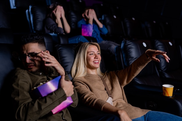 Young smiling woman pointing at large screen while watching action movie with her boyfriend holding box of popcorn