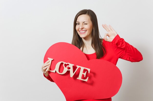 Photo young smiling woman in red clothes showing ok gesture holding big red heart wooden word love on white background. copy space for advertisement. st. valentine's day or international women's day concept