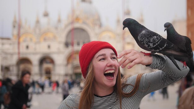 Young smiling woman in red hat stay on the square and feed dove with her hand