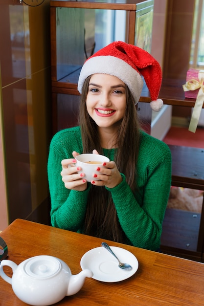 カフェでお茶を飲む赤いサンタ帽子の若い笑顔の女性