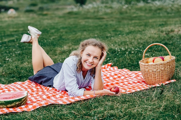 Молодая улыбающаяся женщина отдыхает на открытом воздухе и устраивает пикник, она лежит на одеяле из красных клеток на траве на лугу.
