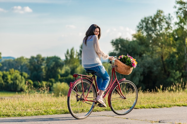 Young smiling woman rides a bicycle with a basket full of flowers in countryside