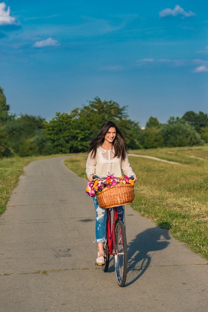 Young smiling woman rides a bicycle with a basket full of flowers in countryside