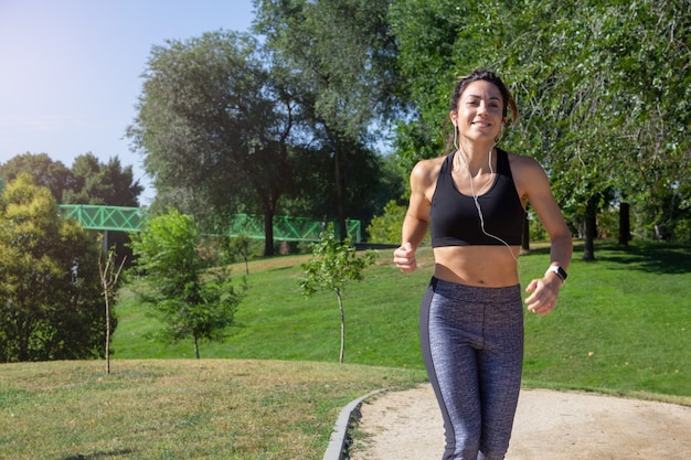 Young smiling woman running through a park on a sunny day while listening to music with headphones