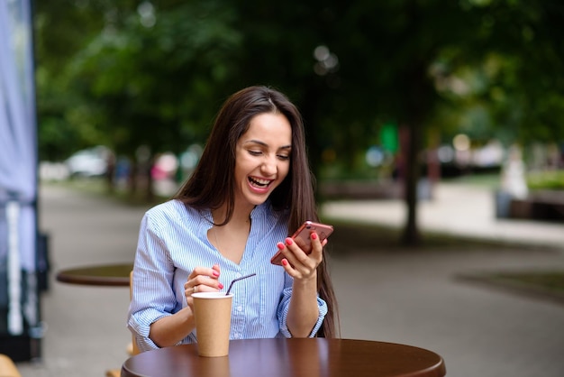 Молодая улыбающаяся женщина делает покупки в Интернете с помощью телефона на открытом воздухе в кафе Красивая модель смотрит на телефон и пьет кофе летом