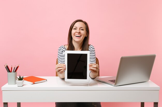 Young smiling woman showing tablet computer with blank empty screen, sit work at white desk with contemporary pc laptop