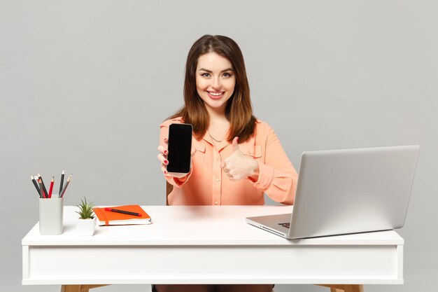 Young smiling woman showing thumb up, holding mobile phone with blank empty screen sit work at desk with pc laptop isolated on gray background. Achievement business career concept. Mock up copy space.