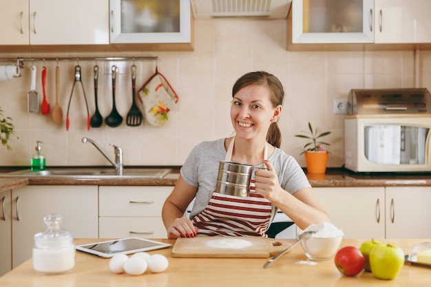 The young smiling woman sifts flour with an iron sieve with tablet on the table in the kitchen. Cooking home. Prepare food.