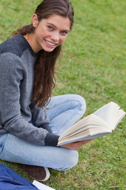 Молодая улыбается женщина сидит скрестив ноги в парке, держа книгу