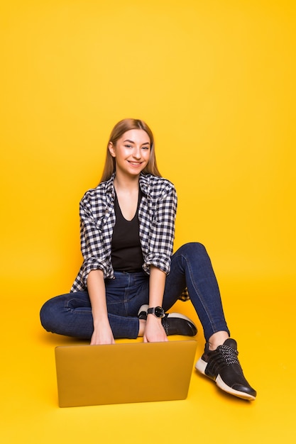 Молодая улыбающаяся женщина сидит на полу с ноутбуком, изолированным на желтой стене