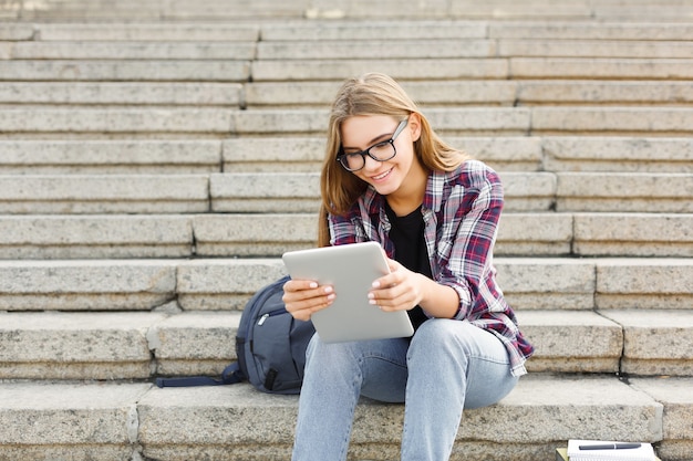 Young smiling woman sitting outdoors on university stairs with digital tablet, typing, surfing internet, preparing for exams. Technology, education and remote working concept, copy space