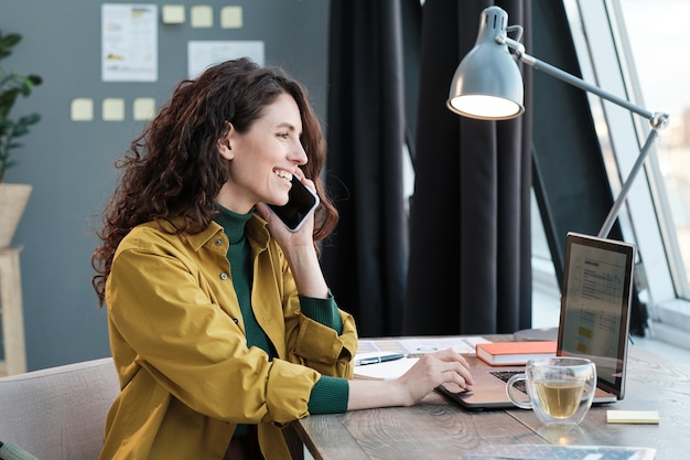 Young smiling woman sitting at the table working on laptop and talking on mobile phone at home