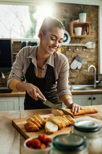 Young smiling woman slicing bread with a knife while preparing food in the kitchen.