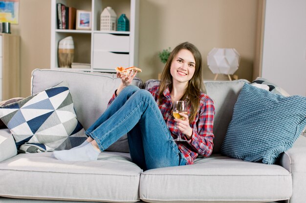 Young smiling woman spending evening at home, sitting on the sofa, drinking wine and eating pizza