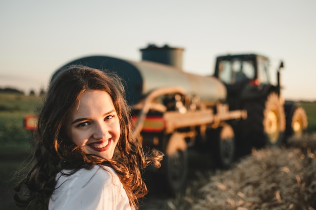 Young smiling woman standing on field on road with tractor and field in sunset background.