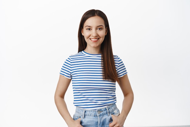 Young smiling woman in striped sea tshirt holding hands in pockets looking ready and happy at camera standing casual against white background