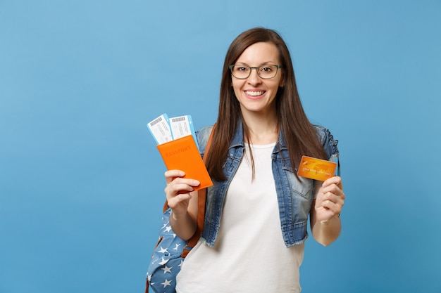 Young smiling woman student in glasses with backpack holding passport boarding pass tickets credit card isolated on blue background. Education in university college abroad. Air travel flight concept.