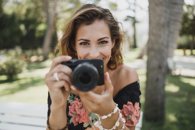 Молодая улыбающаяся женщина фотографирует с помощью своего цифрового фотоаппарата и смотрит в камеру.