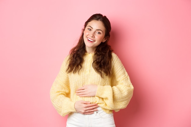 Giovane donna sorridente che tocca il suo stomaco con la faccia felice sollevata che si sente bene dopo aver mangiato yogurt o medicine da crampi dolorosi in piedi contro il muro rosa