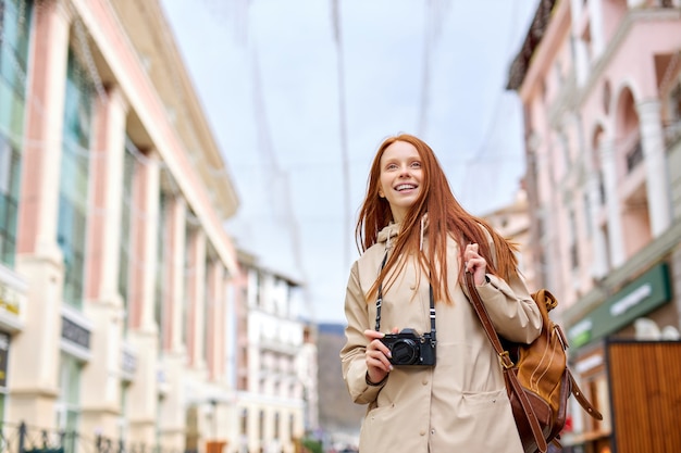 Giovane turista sorridente passeggia per la città in primavera, tiene in mano una fotocamera retrò