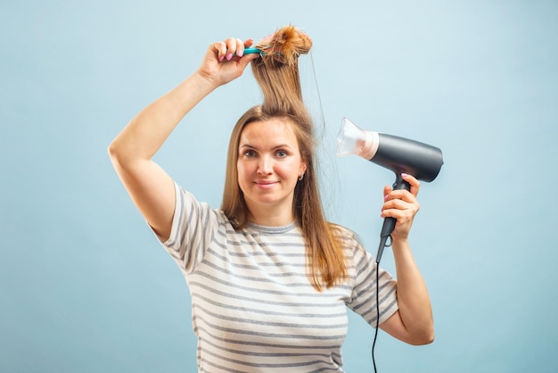 青い背景のヘアケアコンセプトでヘアドライヤーを使っている笑顔の若い女性