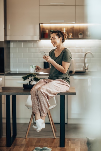 家で朝コーヒーを飲みながらスマートフォンを使う若い笑顔の女性。スマホを使いながらコーヒーを飲む