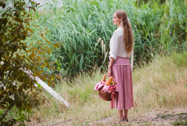 La giovane donna sorridente in abiti vintage in stile retrò tiene un cestino da picnic in mano